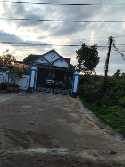 The image depicts a house in Đăk Xú, Vietnam, likely located on the outskirts or in a residential area with adjacent undeveloped land. The house is white with blue trim, featuring a symmetrical design with gabled roofs and decorative symbols. A black metal gate secures the property, which is surrounded by a low wall also painted white with blue accents. A paved driveway leads up to the gate, showing signs of wear. The environment includes greenery, with foliage bordering the driveway and a utility pole partially obscured by vegetation. Overhead, several power lines are visible against a sky with overcast clouds. No people are present in the scene.