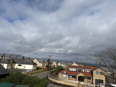 An elevated view depicts a residential neighborhood in Kirklees, United Kingdom, under a partly cloudy sky. A distinct rainbow arches across the middle distance. Numerous houses with light-colored stone facades and dark grey or reddish-brown tiled roofs are visible, arranged along a paved street. In the foreground, lower right, a multi-story stone house with a reddish-brown tiled roof and multiple dark-framed windows includes an integrated garage, beside which a white vehicle is partially parked. Green shrubbery occupies the lower left foreground, and a large, bare deciduous tree stands on the right. In the background, an electrical pylon is visible against distant hills. No people are present, and no actions or interactions are observed.