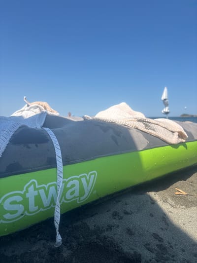 An inflatable kayak, bright green on its lower half and gray on its upper half, lies on a dark sand beach. White and beige towels are draped over the kayak's gray section. A white rope with a patterned stripe hangs from the kayak and touches the sand. The kayak has the word "stway" printed in white letters on its green side. In the background, the blue sky is clear, with a beach umbrella visible in the distance. The overall scene depicts a relaxed beach setting, possibly in Atiya, Bulgaria, with the kayak prepared for or recently used on the water.