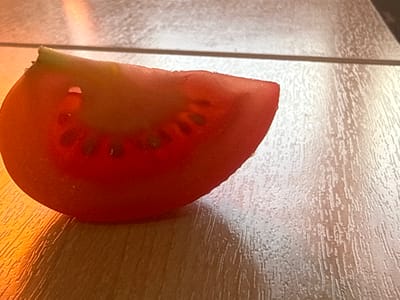 A close-up, low-angle shot shows a single, wedge-shaped slice of red tomato resting on a light-colored, wood-grain surface. The tomato slice, viewed from above, displays its internal structure, including seeds and pulp. The surface it sits on has a visible wood grain pattern and is divided by a thin, dark horizontal line, suggesting it might be a table or countertop. To the left of the tomato, a bright, warm light source casts an orange-yellow glow and creates a soft shadow on the surface. The background above the surface is blurred and has a textured appearance with a vertical stripe pattern, possibly indicating a wall or a different surface. The overall impression is a still life of a fresh tomato slice under dramatic lighting. The image does not contain any information about the city of Sof'ino, Russia.