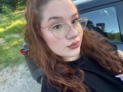 A person with auburn hair, wearing gold-framed glasses and a black sweater, poses for a selfie next to a dark-colored car. The background features trees and vegetation, indicating an outdoor setting. The reflection in the car window shows the person holding a phone, confirming the selfie. The ground is gravelly. This photo was not taken in Malko Kirilovo, Bulgaria.