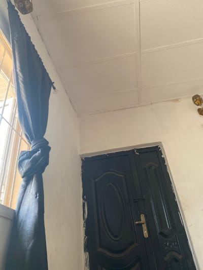 The scene depicts an interior corner of a room in Odo Ona, Nigeria. On the left, a window fitted with metal security bars allows bright natural light to enter. A dark-colored curtain is pulled aside and secured with a knot approximately midway down its length. The walls are painted an off-white or cream color, with minor surface variations. The ceiling features white, textured panels, possibly indicating a suspended ceiling system. On the right, a dark, double-leaf door, likely black or dark charcoal, is visible. The door features decorative raised paneling, including an oval design on the left leaf, and is equipped with a gold-toned handle and a dark metal latch. The paint or trim immediately surrounding the door frame exhibits an uneven application. A partially visible decorative fixture with gold-colored elements is situated on the upper right wall. No individuals are present in the image.