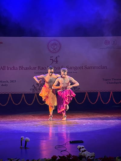The image shows two men performing a dance on a stage in the Tagore Theatre in Chandigarh, India. The stage is lit with purple and blue light, and the floor is made of polished wood. The men are wearing traditional Indian clothing: one is wearing a gold and orange outfit and the other is wearing a pink and gold outfit. The background is a large, white wall with the text "All India Bhaskar Rao Natya & Sangeet Sammelan" in large letters, along with the text "54th", "Unveiling the Soul of Indian Classical Arts", "March, 2025 daily", and "Tagore Theatre Chandigarh". There is a garland of flowers strung along the wall, as well as a small microphone and a black cord lying on the stage floor. The image was taken at coordinates (latitude: 30.733, longitude: 76.789).