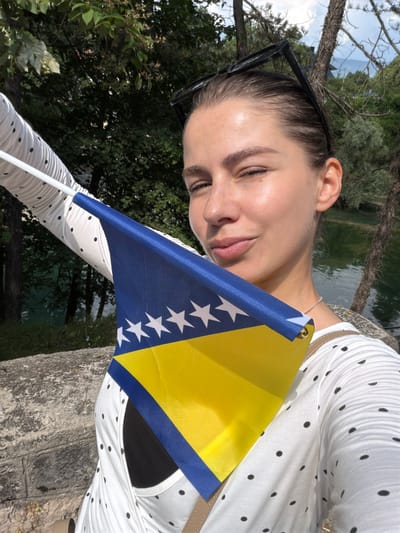 The image shows a young woman holding a small flag of Bosnia and Herzegovina. She is outdoors, with trees and a body of water visible in the background. The sun is shining, suggesting it is daytime. The woman is wearing a white polka dot shirt and sunglasses on her head. Her eyes are closed and she has a gentle smile on her face, conveying a sense of happiness or contentment. The flag she holds is positioned in the foreground, prominently displaying the blue, yellow, and white stars of the Bosnian flag. The overall impression is of a patriotic moment captured in a natural setting. Jajce, Bosnia and Herzegovina is the stated location.