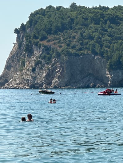 In Đurovići, Montenegro, an image depicts a serene seascape with a prominent rocky cliff face covered in dense green foliage dominating the background. The calm, blue water in the foreground is dotted with several individuals and watercraft. In the middle ground, a small, inflatable boat is anchored, and further to the right, a red and white paddle boat carries several passengers. Closer to the viewer, two people are visible in the water. One appears to be a man wearing sunglasses and a dark shirt, with a second person partially submerged beside him. A cluster of people is also present further out in the water, near the anchored boat. The sky is a clear, pale blue.