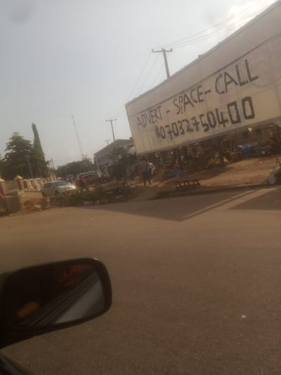 A street scene in Barnawa, Nigeria, captured from a vehicle. A large, light-colored banner with black lettering is prominently displayed, advertising "ADVERT - SPACE - CALL 07032750400." The banner is affixed to a structure that appears to be a market stall or shop, with various goods and tarps visible underneath. In the background, several cars are parked on the side of the road, and people are seen walking. A power pole with wires is also visible. The road surface is asphalt, and the surrounding environment suggests a dusty, outdoor market setting. The image is taken at an angle, with the sky largely obscured by haze or smog. A car's side mirror is in the foreground, reflecting a blurry orange object and a red flower-like shape.