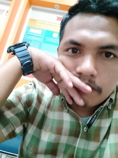 The image shows a close-up, selfie-style shot of a man from the chest up. He is wearing a green and brown plaid shirt over a blue t-shirt. A black digital watch is visible on his left wrist. His right hand is resting on his mouth, partially covering it. The background appears to be an indoor setting with a bulletin board or poster visible behind him, featuring text and graphics in blue and yellow. The lighting is somewhat dim, with a slight reddish tint. The location specified, Meunasahaluedrienlhoksukon Barat, Indonesia, is a town in Aceh, Indonesia, but there are no specific visual cues in the image that confirm this exact location.