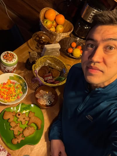 A person in a blue shirt is taking a selfie above a table laden with food. Baskets hold fruit and cookies. There is a bowl of candied fruit, a plate of gingerbread cookies shaped like animals and a witch's hat, and a small dish of gumdrops. A floral-patterned tin sits near the candied fruit. Other containers hold various snacks and items. A coffee maker is also visible.
