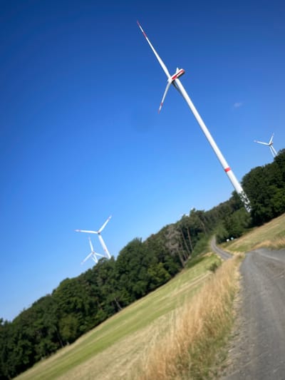 The image captures a rural landscape with wind turbines and a clear blue sky, situated in the vicinity of Berfa, Germany. In the foreground, a dirt road winds its way through a grassy field with dry, golden grasses. The road is flanked by a steep, green grassy slope on the left, which transitions into a dense forest at its upper edge. Several wind turbines are visible in the mid-ground and background, scattered across the landscape and the crest of a hill. The closest wind turbine, in the upper right portion of the image, is tall and white with red accents on its blades and tower. Two other wind turbines are visible further in the distance, appearing smaller and partially obscured by trees. The overall environment suggests a sunny day in a natural, possibly agricultural, setting.