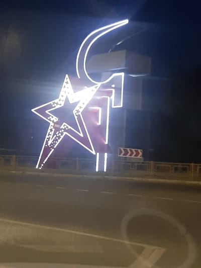 The image shows a nighttime street scene in Shelekhov, Russia, featuring a large illuminated sculpture of a red star with white lights and a sickle. The sculpture is positioned in front of a large concrete structure that appears to be part of a building or monument, also outlined with white lights. The numeral "1" is prominently displayed in white lights to the right of the star. A red and white chevron-style traffic sign is visible on a post to the right of the sculpture, indicating a curve in the road. The sculpture is situated alongside a road with lane markings visible, suggesting it is a roadside installation. A yellow metal fence runs along the side of the road. The overall impression is of an urban environment at night with a striking, possibly symbolic, light installation.
