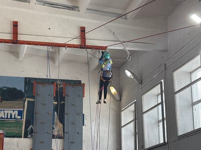 The scene is an indoor facility in Tayshet, Russia, featuring a person suspended mid-air by a system of ropes and climbing equipment. The individual, seen from behind, wears a green helmet, a patterned blue and white top, dark leggings, light athletic shoes, and a multi-colored climbing harness with yellow straps. They are actively engaged with several ropes (blue, red, black, pink, grey) connected to an overhead orange-red metal beam, which is secured to the concrete ceiling via vertical brackets. This beam features multiple attachment points. To the left, a grey panel structure with small protrusions, indicative of a climbing wall, is partially visible, adjacent to a large poster featuring green turf and the partial text 'ATTYL' in blue. The right wall contains multiple rectangular windows fitted with metal mesh screens, allowing external light, complemented by internal fluorescent and dome-shaped light fixtures. The ceiling is composed of exposed concrete beams.