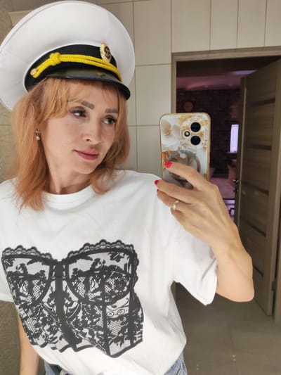 A woman, appearing to be of Caucasian ethnicity with light skin and blonde hair, is taking a selfie in a bathroom. She is wearing a white t-shirt with a black lace corset graphic printed on the front and a white captain's hat with a gold trim and emblem. She is also wearing a ring on her left hand. Her right hand is holding a smartphone with a floral-patterned case. The background shows tiled walls, a doorway leading to another room, and a glimpse of furniture. The image appears to be taken indoors, likely in a residential setting. There is no visible indication of the location being in Kstovo, Russia.
