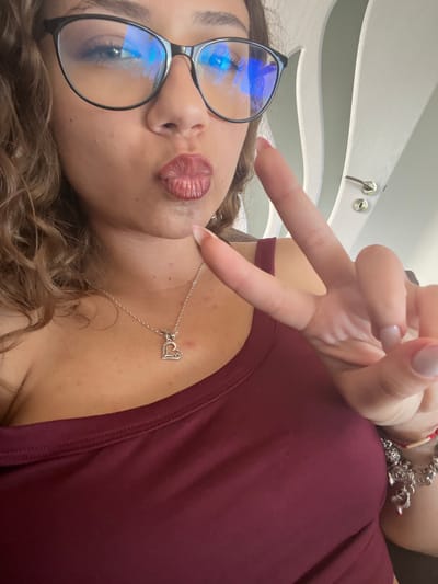 A low-angle self-portrait photograph shows a person with light skin, wearing glasses with black frames and a maroon-colored shirt. They are making a kissing face. The person is holding up their hand in a peace sign. A silver necklace with a heart pendant rests on their neck. A bracelet is on their wrist. The background is a white and grey wall. The location is not determinable from the image.