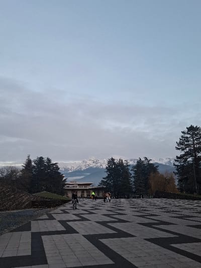 A wide shot of a mountainous landscape features a blue sky with wispy clouds on the left. On the right, a patterned pavement leads to a row of trees and a building. Several people are visible near the building and along the walkway. Snow-capped mountains are seen in the distance. The overall scene suggests a park or outdoor recreational area with natural scenery.