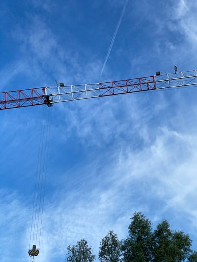 A construction crane with a red and white lattice boom extends across a clear blue sky. Several thin cables hang from the boom, with a hook at the bottom of one set of cables. A faint airplane contrail is visible in the upper right quadrant of the image. In the lower portion of the image, the tops of several green trees are visible. The scene is likely in Lokeren, Belgium, given the context.