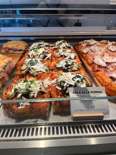 The image displays a vibrant selection of food items, likely in a deli or bakery counter in Dorno, Italy. The primary focus is on two rectangular flatbread pizzas, known as "Pala alla Norma," arranged in a refrigerated glass display case.
The pizza prominently featured in the foreground is generously topped with a rich tomato sauce, slices of eggplant, fresh green herbs (likely basil), and a generous scattering of shredded white cheese, which the label identifies as salted ricotta. This pizza has been cut into several individual portions, indicating it's ready for sale by the slice. Above it, another rectangular pizza is visible, adorned with what appears to be thin slices of cured meat or cheese, dusted with white powder, possibly grated hard cheese. Below these main items, other food products, including sandwiches or further flatbreads, can be partially seen.
A white price tag with black text is clearly visible, describing the foreground pizza. It reads: "PALA ALLA NORMA," followed by "Pizza alla pala con salsa di pomodoro, melanzane e ricotta salata" (Flatbread pizza with tomato sauce, eggplant, and salted ricotta). Below this, "and salted ricotta cheese" provides an English translation. The price listed is "€ 7,90." Further down, text in Italian and English states: "Ingredienti e allergeni su Cartelli Ingredienti. Prodotto con base congelata all'origine." and "Ingredients and allergens on Ingredients Signs. Product with base from frozen at source."
The scene suggests a casual eatery or food market, with the food presented appealingly for customers. The metallic elements of the display case, including a grated vent system on the right, add to the commercial setting. The lighting is bright, illuminating the food clearly.