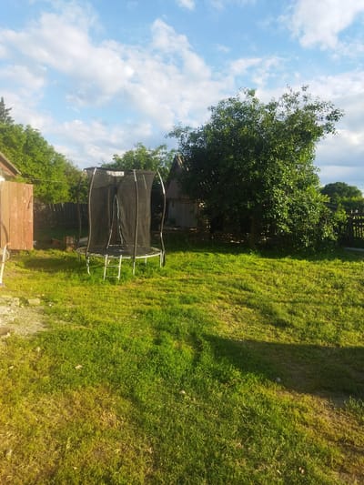 The image depicts a backyard scene in Sosnivka, Ukraine, under a partly cloudy blue sky. In the foreground, a well-maintained green lawn is visible, with patches of sunlight and shadow. A trampoline with a black safety net and a silver frame stands in the middle ground, accompanied by a silver ladder. To the left of the trampoline, a weathered wooden fence and part of a building are present. Further in the background, dense green trees and shrubbery are seen, as well as another rustic wooden structure with a corrugated metal roof. A wooden fence encloses the right side of the yard. The overall impression is a peaceful, rural domestic setting. There are no people or animals clearly visible in the frame.