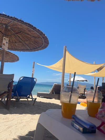This is a ground-level, eye-level shot of a beach scene at Ofrynio Beach, Greece. In the foreground, a white plastic table holds two clear plastic cups with yellow drinks and black straws, along with a blue vape pen and its packaging. In the mid-ground, beach chairs and sun loungers are arranged on the sand. A blue beach chair is prominently visible, with a beige chair to its left. Further back, a person is visible lying on a sun lounger. The background features the turquoise sea, a calm horizon with distant landmasses, and a clear blue sky. Large thatched umbrellas and yellow sunshades provide shade on the beach. The overall atmosphere is one of relaxation and summer leisure.