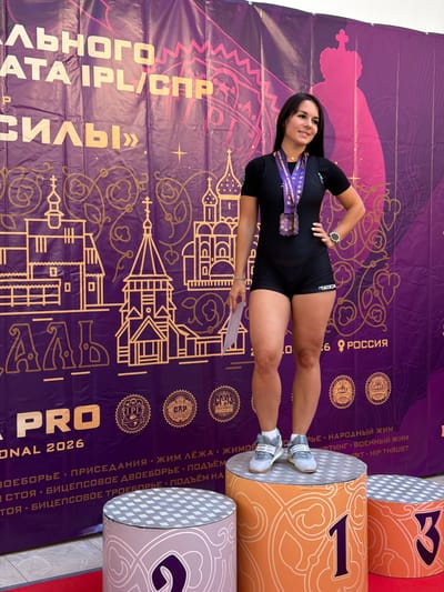 A woman stands on a podium in front of a purple banner. The banner has gold lettering and depicts stylized Orthodox churches and crowns. The woman is wearing a black athletic outfit, a medal around her neck, and white athletic shoes. She is holding a piece of paper in her left hand and has her right hand on her hip. Her gaze is directed to her left. The podium she is standing on is cylindrical and decorated with an orange pattern. The number "1" is prominently displayed on its side. To her right, another podium, also cylindrical and orange with a checkered top, displays the number "3". To her left, a portion of a third podium is visible, decorated in pink and white. The background banner includes text in Russian, including "IPL/СПР", "РОССИЯ", and "НАРОДНЫЙ ЖИМ". The event appears to be a powerlifting competition, possibly related to the IPL (International Powerlifting League) and SPR (Soyuz Pauerliftingov Rossii). This event is implied to be taking place in Suzdal, Russia, due to the presence of Russian text and the stylistic elements on the banner, which evoke Russian cultural motifs.