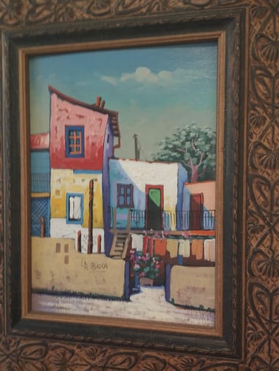 This is a framed painting depicting a colorful, slightly abstract architectural scene of multi-story buildings with vibrant facades in shades of pink, red, yellow, and blue. The buildings have various windows and doors, some with wrought-iron balconies. Clothes are hanging on a line in front of some of the buildings, adding a domestic element. A tree with green foliage and a pinkish trunk is visible in the background. The sky is light blue with a few white clouds. The painting is signed "LA BOCA" in the lower center. The frame is ornate and appears to be made of wood with carved patterns. The image does not contain any information or visual cues that suggest a specific location in Pardes Hana-Karkur, Israel.