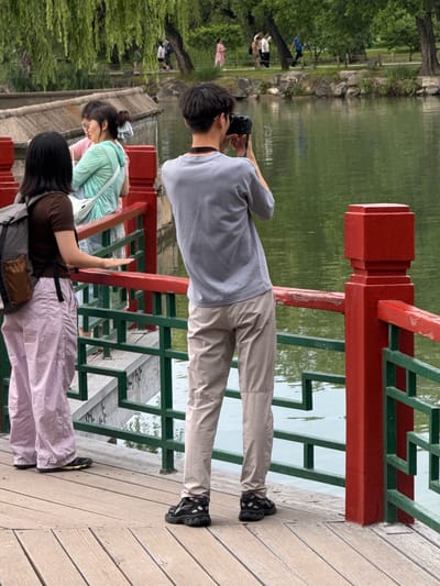 The image depicts a scene on a wooden bridge adjacent to a body of water within a park in Haidian District, China.
On the bridge, three individuals are visible in the foreground. On the right, a young male, viewed from behind, wears a light gray short-sleeved t-shirt, light khaki trousers, and black athletic shoes. He holds a black digital camera to his eye, aiming it towards the left, indicating he is photographing the scene. To his left and slightly forward, a young female, also viewed from behind, wears a dark brown short-sleeved t-shirt, light purple wide-legged trousers, and dark athletic shoes with yellow accents. A gray and brown backpack rests on her left shoulder, and her right hand is placed on the red railing of the bridge. Further left, partially obscured by the foreground female and the railing, a third young female faces left, wearing glasses, a light mint green long-sleeved top, and a white cross-body bag.
The bridge features a wooden plank walkway and a distinctive railing system composed of prominent red horizontal top bars and vertical posts, interconnected with a geometric pattern of green bars below. The red paint on the railing shows noticeable wear.
The bridge overlooks a calm body of water with a greenish hue, reflecting the surrounding environment. The opposite bank of the water is densely populated with lush green trees, including weeping willows whose branches extend towards the water. A stone embankment or retaining wall lines the far bank, behind which a walking path is visible. Several indistinct figures are seen walking along this path in the background, one appearing to wear a light pink dress. The overall environment suggests a recreational park setting.