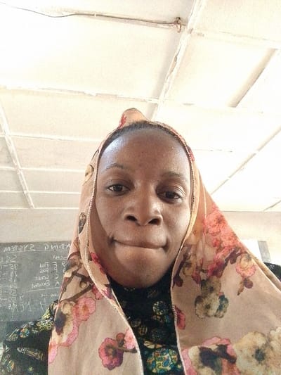 This image depicts a close-up, head-and-shoulders view of an adult female in an indoor setting within Barnawa, Nigeria. The woman, with a dark complexion, is centered in the frame, looking directly at the camera with a neutral expression and slightly pursed lips. She wears a light beige head covering, likely a hijab, adorned with a prominent floral pattern in shades of pink, orange, and green. A dark, patterned garment is visible underneath the head covering. The background on the left reveals a dark chalkboard with white, inverted writing, including partial words such as "SYSTEMS" and "ADING," and various numerals, suggesting a classroom environment. The upper background features a light-colored ceiling with a grid of square tiles, exhibiting some visible wear and a thin dark wire running across it.