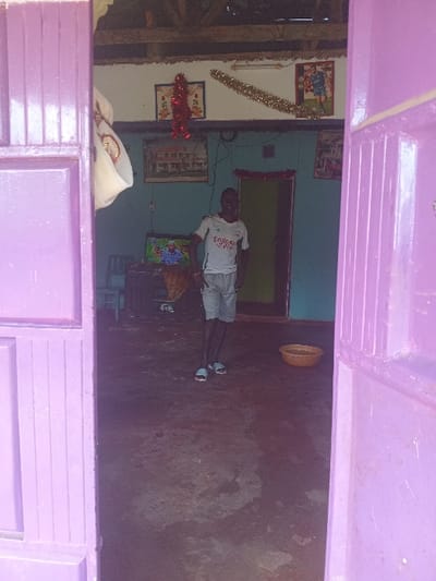 This image captures an indoor scene, viewed through a purple, possibly shuttered, doorway or window frame. The primary subject is a young person, likely a boy, standing centrally within a modest room. He is wearing a white t-shirt with the text "EMIRATES FLY BETTER" emblazoned on the front, dark shorts, and light-colored flip-flops. He looks directly at the camera with a neutral expression.
The room features walls of differing colors: a light blue or teal wall on the left, adorned with several framed pictures and festive decorations, including a vertical golden garland and a horizontal red tinsel piece. On the right, the wall is a darker, earthy tone, possibly concrete or mud, showing some patches of wear. A dark doorway or door is visible in the background between the two walls.
A small television is placed on a low stand near the center of the room, to the right of the boy. On the dark floor, which appears uneven, a large, round, light brown woven basket or bowl rests near the back wall on the right. In the bottom left corner of the internal room, partially obscured by the foreground frame, a white piece of fabric is draped, containing small, reflective objects, possibly jewelry. The ceiling, visible in the upper left, shows exposed wooden beams, suggesting a simple construction.
The overall impression is that of a humble dwelling, possibly in Manda-Shivanga ward, Kenya, consistent with the provided location context. The lighting is somewhat dim, suggesting an indoor setting without much natural light, or possibly evening. There is no specific activity taking place beyond the person posing for the photograph.