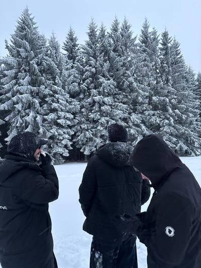 In Le Hohwald, France, three individuals are observed outdoors in a dense, snow-covered forest environment. The ground is uniformly blanketed with fresh snow, and numerous coniferous trees in the background are heavily laden with snow or hoarfrost. The sky is overcast and grey. All three individuals are dressed in dark winter jackets and attire, with visible snowflakes accumulating on their clothing, suggesting ongoing snowfall.
The person on the left is seen in profile, wearing a dark hooded jacket and a baseball cap beneath the hood. They are bringing a white object, consistent with a tissue or handkerchief, to their face with a gloved right hand. The middle individual is viewed from the back, wearing a dark puffer jacket with the hood down, revealing a dark beanie. The person on the right is seen from a three-quarter perspective, wearing a dark hooded jacket with the hood up. This individual is looking down and holding a dark, rectangular electronic device, likely a smartphone, in gloved hands. A circular patch on their left sleeve displays a wolf and moon graphic with the text "LE RÊVE DES LOUPS." The individuals are standing separately, without direct interaction, and are generally oriented towards the forest.