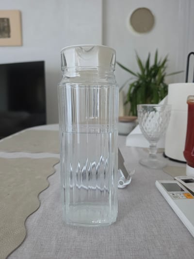 A clear glass pitcher filled with water stands on a light gray textured tablecloth. The pitcher has vertical ribbed details and a white lid. To the right of the pitcher, a clear glass goblet with a patterned design sits on the tablecloth. Further to the right, a white roll of paper towels and a red bottle, possibly containing ketchup, are visible. In the background, a green plant in a pot, a framed picture on a light-colored wall, and a dark television screen are discernible. A white remote control is placed on the tablecloth in the foreground. The scene appears to be indoors, possibly in a dining area or living room. The image does not provide any specific clues to its location in Almenara, Spain.