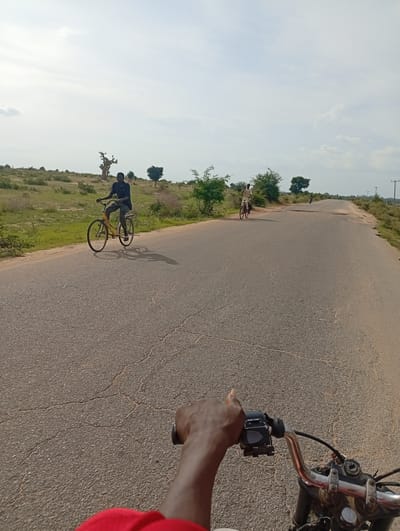 In Shuwa, Nigeria, this image captures a view from a motorcycle moving along an asphalt road. Two individuals on bicycles are visible in the distance on the same road. The landscape bordering the road is characterized by dry grass, scattered bushes, and a few trees, including a distinctive baobab tree. The sky is partly cloudy, with the sun casting shadows on the road. The focus is on the road ahead and the handlebars of the motorcycle, with a hand on the right side giving a thumbs-up gesture.