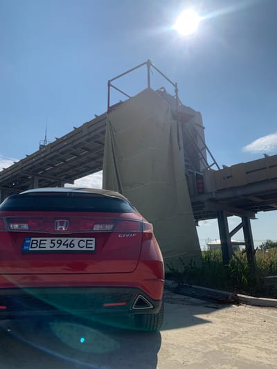 A red Honda Civic is parked outdoors on a paved surface. The car is facing away from the camera, and the license plate is visible. The license plate reads "BE 5946 CE" with a Ukrainian flag symbol on the left. The car is positioned in front of a large, unfinished or industrial structure, possibly a construction site. This structure appears to be made of metal beams and covered in a large tarp. The sun is shining brightly in the sky, creating lens flare in the image. The sky is blue with some wispy clouds. The overall scene suggests a sunny day at an industrial or construction location in Pivdennoukrainsk, Ukraine.