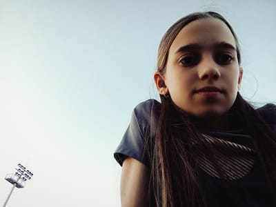 The image shows a young person with long brown hair, wearing a dark t-shirt with a striped pattern on the collar. The person is viewed from a low angle against a bright, clear sky. To the left of the person, in the distance, is a tall light pole with multiple lamps. This scene appears to be an outdoor sports field, likely at a stadium or park. The environment suggests it is daylight. The young person is looking directly at the camera. The specific location in Radzymin, Poland, cannot be determined from the visual information in the image alone, as there are no identifying landmarks or specific architectural features that would pinpoint a precise location within the city. However, the presence of a sports field light suggests a public recreational area.