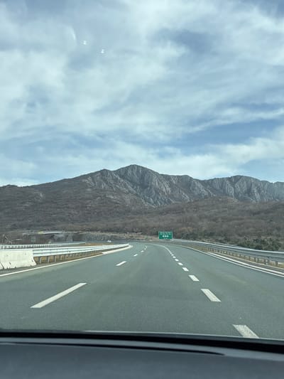 This image presents a forensic view of a highway segment near Pelev Brijeg, Montenegro, observed from within a vehicle. The asphalt highway features two lanes of travel in the foreground, demarcated by a solid white line on the left shoulder and dashed white lane separators, curving gently to the right. White concrete barriers line the left side, transitioning to metal guardrails further along, with additional metal guardrails lining the right side.
In the mid-ground, a green overhead road sign is visible, displaying destinations such as "Bioče," "Veternik," and "Podgorica" with associated distances, in both Cyrillic and Latin script. A partial bridge or overpass structure is visible to the left, beyond the concrete barriers. Minimal vehicle traffic, appearing as small white forms, is discernible in the distant lanes.
The background is dominated by a large, rugged mountain range characterized by prominent grey rock formations and slopes covered in sparse, bare deciduous vegetation, indicating a late autumn or winter season. The sky above is partly cloudy, exhibiting a mix of light blue and grey cloud formations. The scene is illuminated by natural daylight.