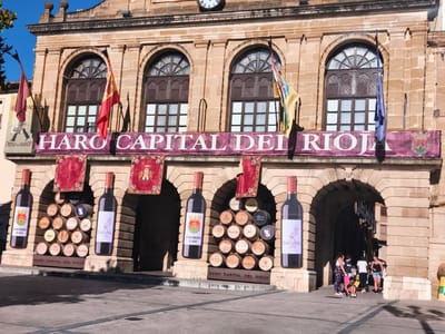 The image captures a sunny day in Haro, Spain, specifically at the iconic Haro Capital del Rioja building. The facade of the building is made of warm, tan-colored stone with arched windows and decorative stonework. A large banner across the building reads "HARO CAPITAL DEL RIOJA" in prominent white lettering against a deep red background. Several flags are displayed, including the Spanish flag and other regional banners.
In front of the building, two oversized wine bottles are displayed as decorations, one on each side. Between these bottles, a collection of wine barrels is stacked in a pyramid shape. Each barrel appears to be labeled with names of wineries or brands, suggesting the area's rich wine-making heritage. The ground in front of the building is a paved public square.
Several people are visible in the scene. On the right side of the image, a group of individuals is gathered near the arched entrance of the building. Some are standing and conversing, while others appear to be walking or pushing a stroller. Their attire suggests casual daytime activity. The overall atmosphere is festive and celebratory, likely tied to a local event or the promotion of the region's wine culture.