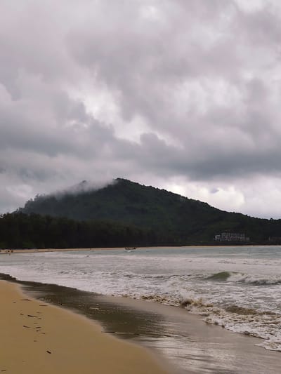 This is a photograph of a beach in Sakhu, Thailand, under an overcast sky. The foreground shows a wet, sandy beach with scattered debris. The middle ground features the ocean with moderate waves breaking on the shore, creating white foam. In the distance, a dark green, forested hill rises, with clouds clinging to its peak. A small cluster of white buildings is visible on the lower slope of the hill on the right side of the frame. A narrow, dark boat is positioned on the water near the shoreline. Several figures, appearing as small dots, are scattered along the beach and in the shallow water. The overall atmosphere is muted, consistent with a cloudy day.