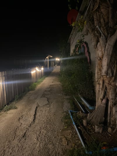 The image depicts a narrow pathway illuminated by string lights in Baan Don Sai, Thailand, at night. The pathway is bordered by vegetation and a cracked, uneven surface. The left side of the path is dark, with a glimpse of a structure with string lights. Overhead, there are tree roots, exposed pipes, and debris. In the top-left corner, there's a glimpse of a red object mounted on a wall with graffiti art. A broom leans against the wall above the path.