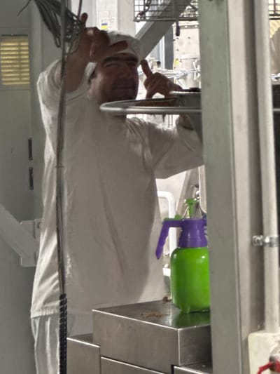 A man wearing a white chef's hat and uniform is standing in a brightly lit room. He is holding his hands up in a gesture that resembles playing horns or a victory sign. In the foreground, a green spray bottle with a purple nozzle sits on a metallic surface. The background appears to be an industrial or laboratory setting with various equipment and structures. The image has a slightly grainy quality.