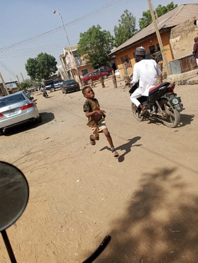 The image depicts an unpaved, dusty street scene in Gama, Nigeria. In the foreground, a male child, estimated to be 5-8 years old, is captured mid-stride, running from the right towards the left. He wears a dark t-shirt with a graphic, light-colored shorts, and sandals, displaying an intense facial expression. His shadow extends to his right. A dark, circular object, consistent with a vehicle mirror, is partially visible in the lower-left quadrant.
In the mid-ground, a male individual in a white traditional garment and white cap is riding a black motorcycle, moving from left to right, away from the child.
The background features various buildings, including a multi-story structure on the left and single-story buildings with weathered, light brown/orange walls on the right. A silver sedan (Honda Accord) is parked on the left, and further back, a blue and white police vehicle is visible, along with other parked cars (including a red sedan) and another distant motorcycle. A street light pole stands centrally. Green trees are interspersed throughout the background. The scene is under clear skies with strong sunlight, indicated by pronounced shadows. Wooden stakes are visible along the right side of the road.