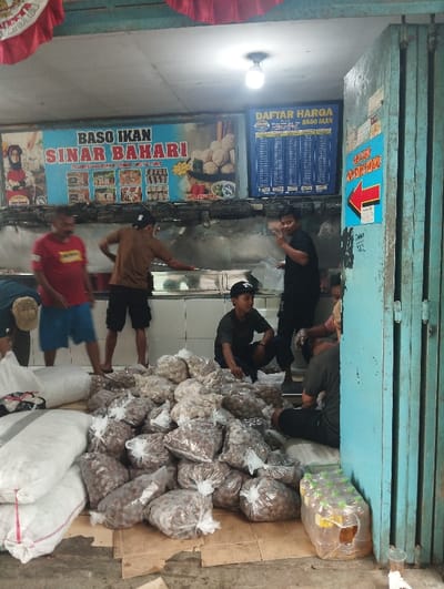 This image captures a bustling indoor commercial space in Baleendah, Indonesia, likely a production or distribution point for meatballs. The scene is dominated by several men, appearing to be workers, engaged in handling large quantities of food.

In the foreground and mid-ground, numerous clear plastic bags filled with meatballs (baso) are piled high on cardboard sheets on the floor, alongside several large white woven sacks, possibly containing more meatballs or raw ingredients. A few men are squatting or standing around these piles, suggesting they are sorting, packing, or moving the products. In the background, three men stand behind a counter or work surface. One man on the right, wearing a black shirt, looks towards the camera and makes a hand gesture.

Overhead, a bright fluorescent light illuminates the scene, indicating an indoor setting during what appears to be daytime. The walls in the background feature tiled surfaces, common in food preparation areas. To the right, a folded blue metal accordion-style door or shutter marks the entrance or exit.

Visible text includes a large banner across the back wall that reads "BASO IKAN SINAR BAHARI" (Fish Meatballs Sinar Bahari), featuring images of various products. Next to this, a blue sign displays a "DAFTAR HARGA BASO IKAN" (Price List Fish Meatballs). A smaller, bright blue sign with a red arrow pointing left is affixed to the blue shutter, with some unreadable Indonesian text. The overall impression is that of a local, high-volume operation focused on the production and sale of fish meatballs.