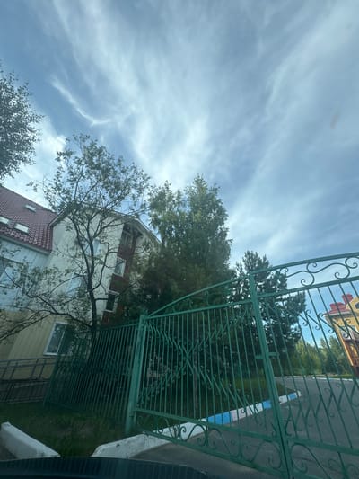 A low-angle, outdoor, eye-level shot shows a residential building and a green metal fence. The building is multi-story with a red-tiled roof and light-colored walls, featuring several windows. A tree with sparse foliage stands in front of the building. The green metal fence, with decorative scrolling patterns, runs across the foreground and right side of the image. Beyond the fence, a paved area with painted lines and more trees is visible. The sky above is partly cloudy with wispy, white clouds against a light blue background. The scene appears to be in a residential area in the city of Megion, Russia.