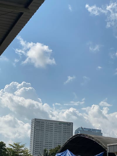 The image captures a daytime scene in Gambut Barat, Indonesia, dominated by a bright blue sky filled with fluffy white clouds. In the foreground, a portion of a grey, corrugated metal roof overhangs the frame from the top left, casting a shadow. Below and to the right, two modern buildings rise against the sky. The taller building on the left is a light-colored, multi-story structure with numerous windows, appearing to be an office building or hotel. A sign with green text is visible near its top. To the right of this building is a shorter, glass-fronted building with a prominent rooftop sign. In the bottom portion of the image, a large, dark, curved canopy or tent structure partially obscures the view of trees and other objects below, with a blue tarp visible underneath it. The overall impression is an urban landscape under a clear, sunny sky with scattered clouds.