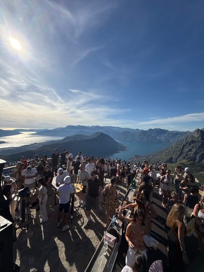An outdoor party is taking place on a high vantage point overlooking a bay and mountains. The sun is shining brightly in a blue sky with wispy clouds. A crowd of people, dressed in casual summer attire, are gathered on a stone-tiled terrace. Some are standing around tables, others are conversing, and some appear to be dancing. Beverages and some bottles are visible on the tables. The background shows a scenic view of a bay dotted with boats, surrounded by lush green mountains. A town or village is nestled along the coastline. The overall impression is a lively social gathering in a picturesque natural setting. The location is identified as being in the vicinity of Špiljari, Montenegro.