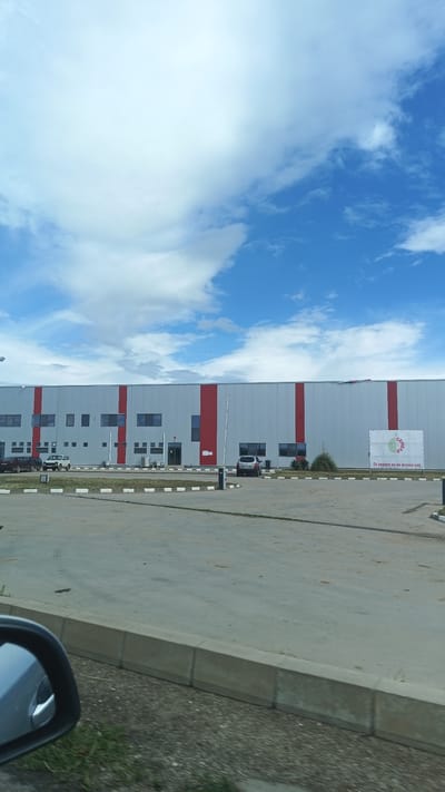 The image shows a long, gray industrial building with red vertical stripes. It is located in Градина, Bulgaria. The building has a number of windows and doors. A sign with the word "семейство" is on the right side of the building. There is a paved parking lot in front of the building with some parked cars. The sky is blue with white clouds. The photograph appears to have been taken from inside a vehicle, with the side mirror visible in the lower left corner of the frame. A concrete curb and grassy area are visible at the bottom of the image.