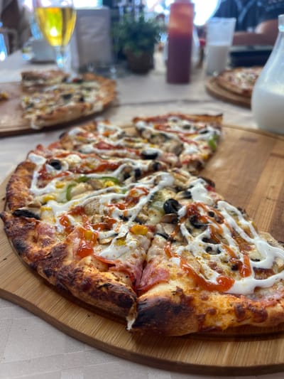 In the foreground of the image, a large, partially eaten pizza is presented on a round wooden board. The pizza is topped with various ingredients including mushrooms, corn, green bell peppers, ham, and black olives. It is drizzled with a white sauce, possibly mayonnaise or a creamy dressing, and a red sauce, likely ketchup or tomato sauce. The pizza crust is visibly charred in several spots, indicating it was baked at a high temperature.

Behind the main pizza, another pizza can be seen on a similar wooden board. Further in the background, the scene appears to be a restaurant or outdoor dining area. A glass of light-colored beverage, likely beer, is visible on the left. A red bottle, possibly ketchup, and a glass of milk are also present. A white pitcher, also possibly containing milk, is on the right side of the frame. The environment suggests a casual meal setting.

The image was taken in Kranevo, Bulgaria.