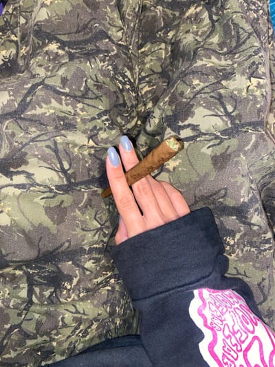A close-up image captures a person's hand holding a cigar or blunt. The hand has light blue nail polish on the fingernails and emerges from the cuff of a black long-sleeved garment, likely a sweatshirt. Part of a white and pink graphic design is visible on the black garment, featuring stylized text that appears to include "WOOFEAR" or similar letters. The cigar/blunt is rolled in a brown wrapper, with green, plant-like material visible at the tip. The background is a camouflage-patterned fabric in shades of green, brown, and dark grey. The scene suggests a casual, indoor setting in Monte Grande, Argentina.