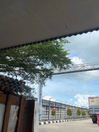 The image depicts an outdoor scene in the city of Gambut Barat, Indonesia. In the foreground, a portion of a gray, corrugated roof overhang is visible, with a glimpse of lush green foliage from a tree beneath it. To the left, stacked egg cartons and wooden crates are partially visible. The midground features a concrete wall topped with barbed wire, and a line of small, ornamental trees with red-tipped foliage. Beyond the wall, a white pedestrian bridge with metal railings spans across the sky. The sky itself is a mix of blue and white clouds. On the right side of the image, a truck with a red trailer is parked. The ground is paved with asphalt, and a black and white striped border runs along the edge of the walkway. The overall impression is one of an industrial or logistical area.