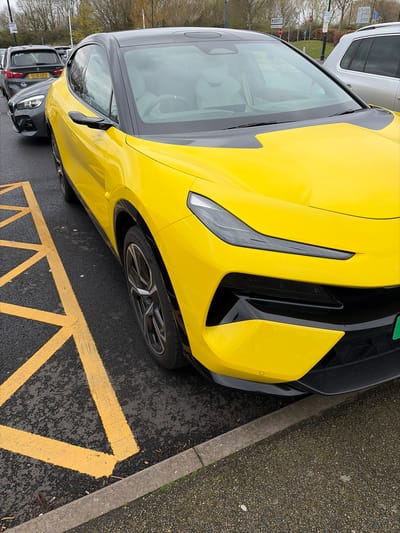 The image shows a close-up of the front portion of a yellow sports car parked in a marked parking space with yellow diagonal lines on a dark asphalt surface. A concrete curb is adjacent to the parking space. Other vehicles are partially visible in the background, including a gray SUV and other cars parked in a row. The car has a black roof and a black front grill. The parking space appears to be located in an outdoor parking lot, possibly in Braintree, United Kingdom, given the prompt. There are no people visible in the image.