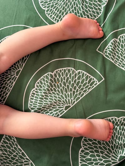 The image shows the lower legs and feet of a child lying on a green bedsheet with a white floral fan pattern. The child's skin is light-skinned. One foot is visible from the top, showing toes and the arch of the foot. The other foot is visible from the sole, showing the heel and the ball of the foot. The child's legs are bent. There is a small orange fleck on the bedsheet near the child's legs. The image is taken from a top-down perspective. There is no discernible information about the location in Lopatino, Russia.