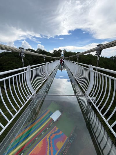 The image depicts a glass-bottomed suspension bridge in Lam Vien - Da Lat, Vietnam. The bridge is constructed with white metal railings and thick glass panels that allow a view of the forested area below. Several people are walking on the bridge, their reflections visible in the glass. The sky is filled with white, fluffy clouds against a blue backdrop, and lush green trees surround the bridge. The glass panels of the bridge also feature a colorful, geometric pattern beneath the surface, possibly a decorative element or part of the underlying structure.