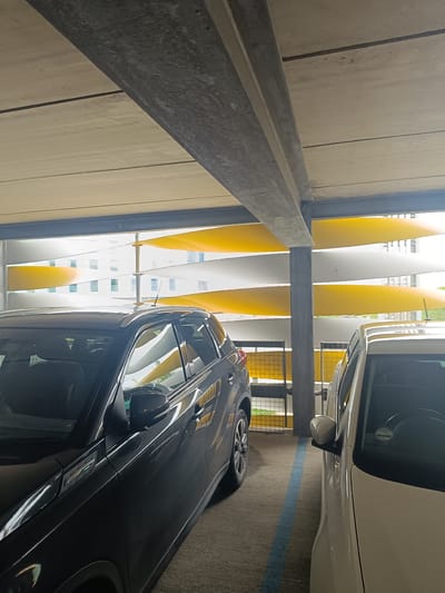 This is a photograph taken inside a multi-story car park. The image shows the interior of a car park with concrete beams and a yellow and white slatted facade visible in the background. Two cars are parked side-by-side, one dark-colored SUV on the left and a lighter-colored car on the right. A blue parking line is visible on the ground between the two vehicles. The lighting suggests it is daytime, with natural light entering the car park from the open facade. The location is identified as a car park in Cambridge, United Kingdom.